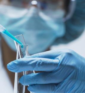 A lab technician wearing blue gloves pipettes blue liquid into a test tube