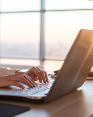 Woman on laptop A businesswoman types on a laptop as the sun sets through the large windows behind her