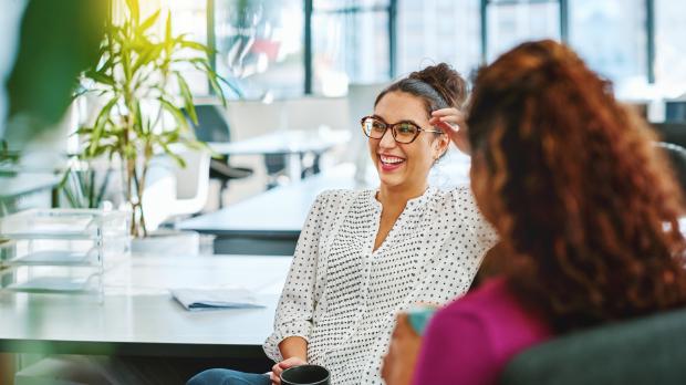 A woman sits with a cup of tea, chatting with her colleague and smiling in a bright modern office