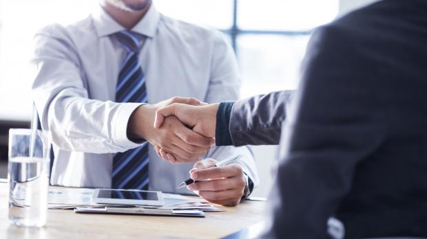 A man in a shirt and tie sitting at a table shakes the hand of a man in a business suit